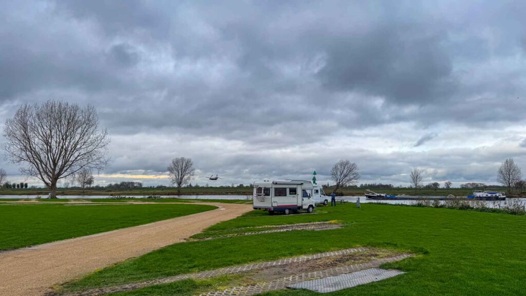 Camper op een camperplaats bij Heusden met op de achtergrond een passerende Chinook helikopter en een boot